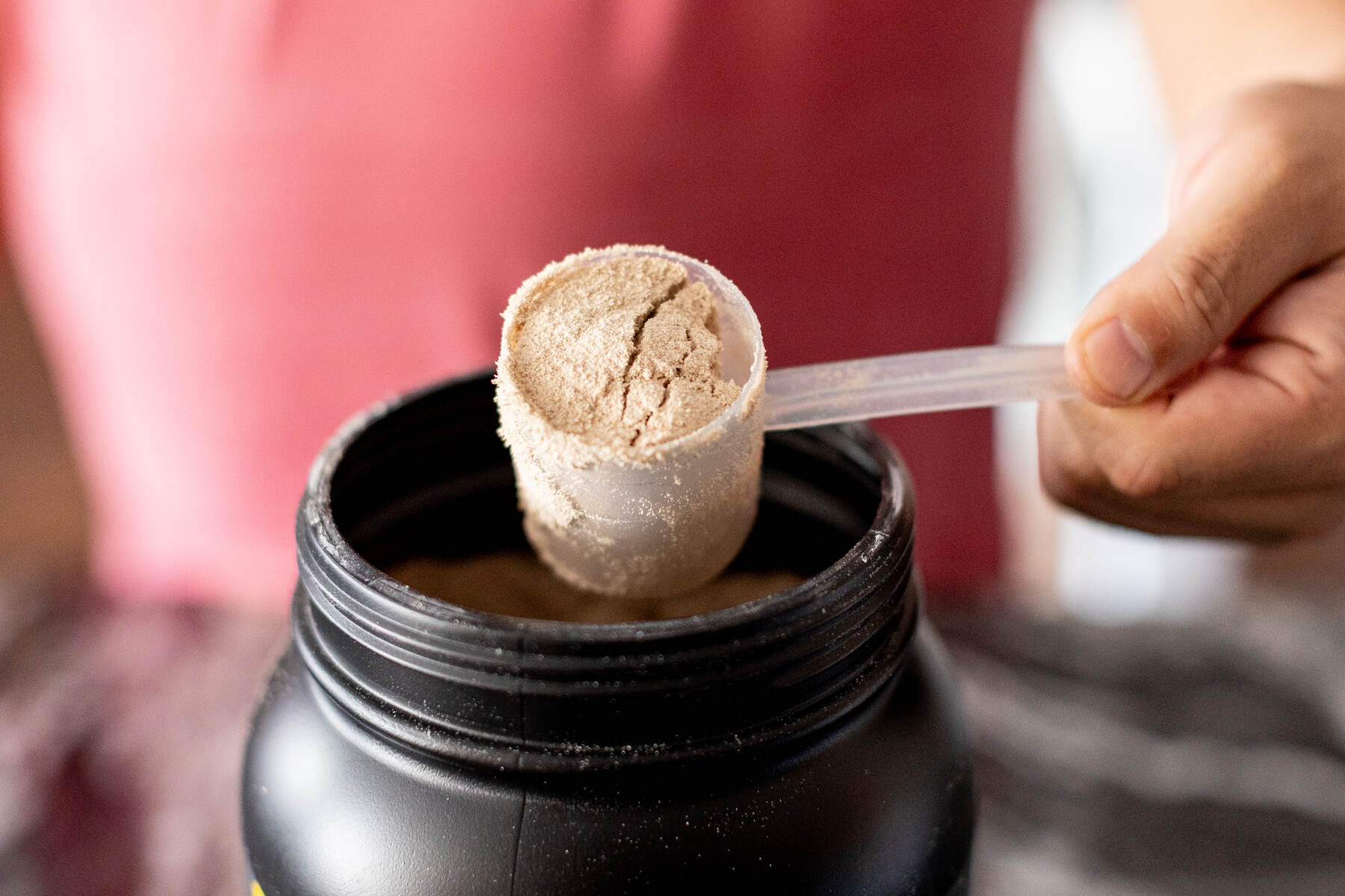 Man holding a scoop of chocolate protein powder above the black container on a kitchen countertop