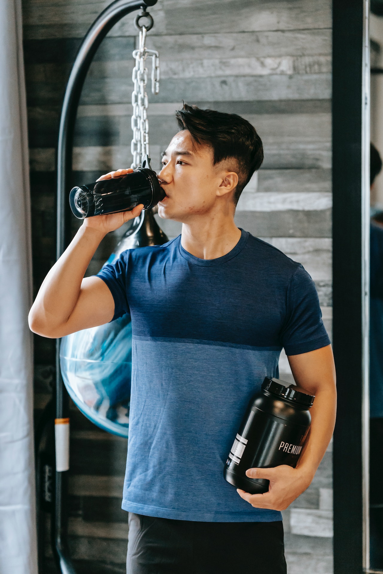 A man wearing a blue shirt is drinking from a black tumbler while holding a black jar of pre-workout supplements inside the gym