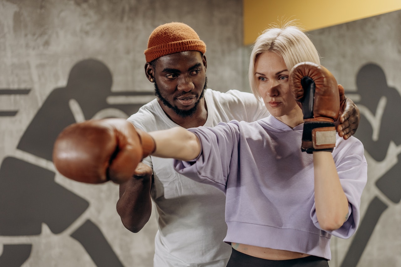 A man wearing a white shirt is teaching boxing to a girl wearing a purple hoodie and brown boxing gloves inside a gym