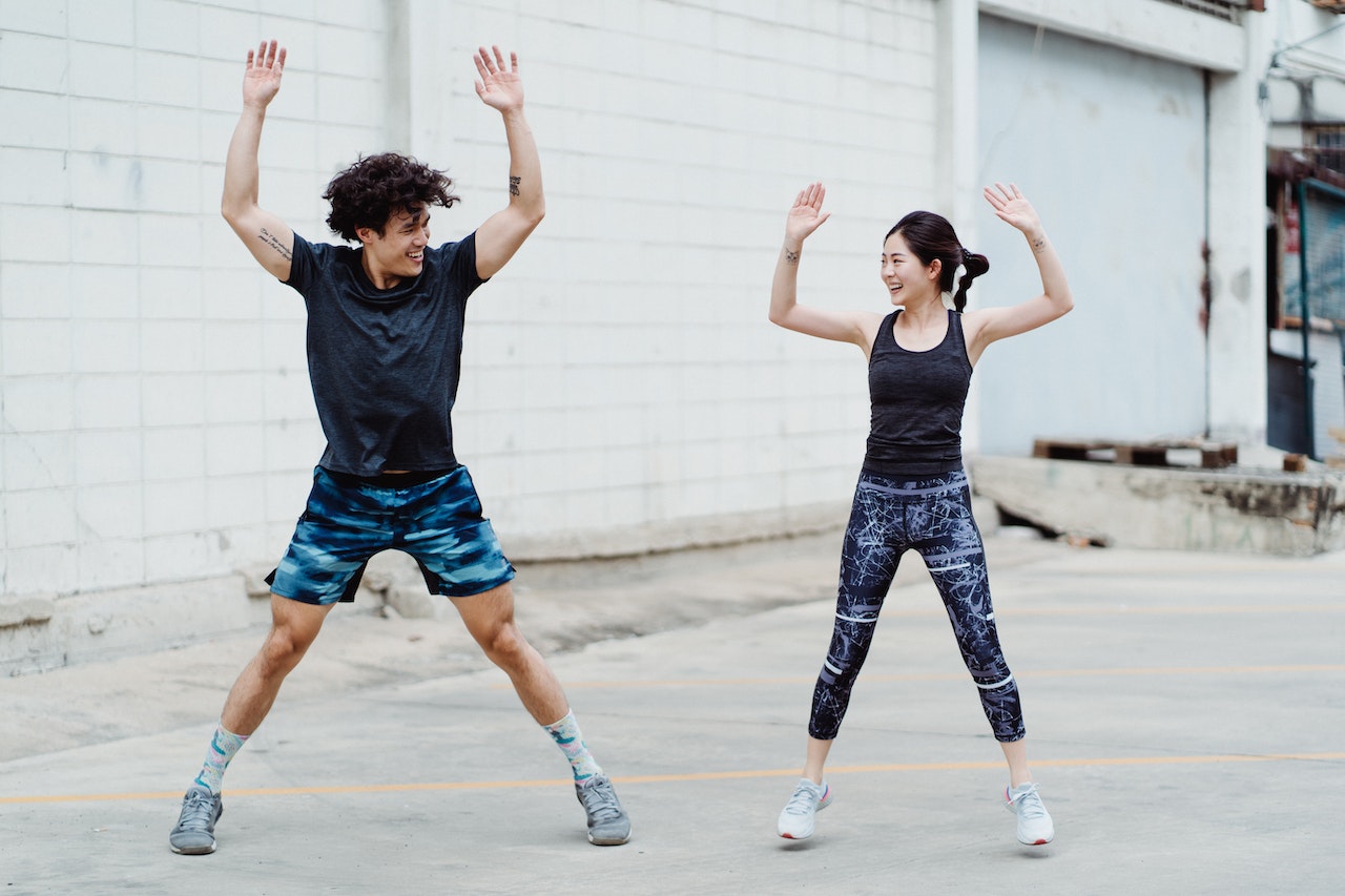 A man wearing a black shirt and blue shorts is doing jumping jacks beside a girl wearing a black tank top and printer leggings