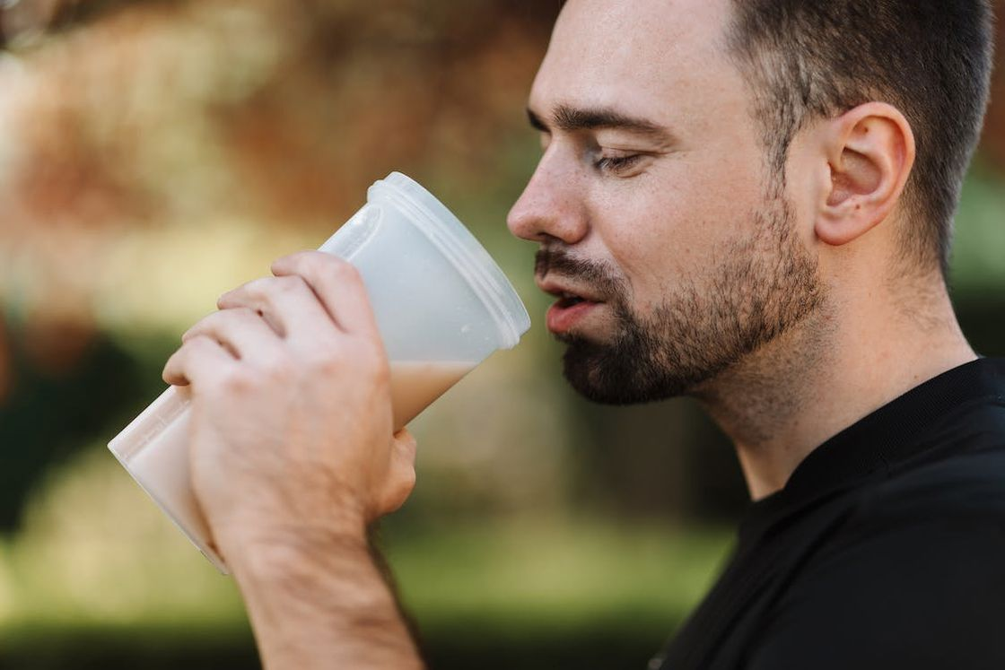 Man drinking his protein shake from a clear plastic tumbler during his break while exercising outdoors