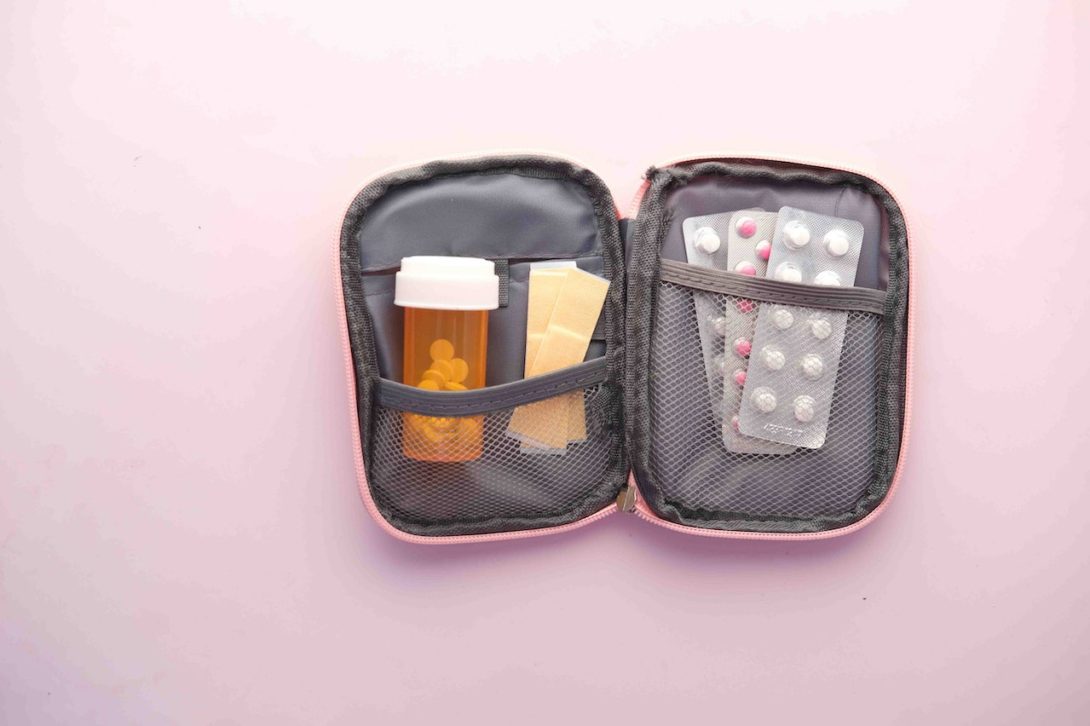 A gray and pink colored medicine bag with an orange medicine bottle, yellow adhesive bandages, and supplements on top of a pink surface