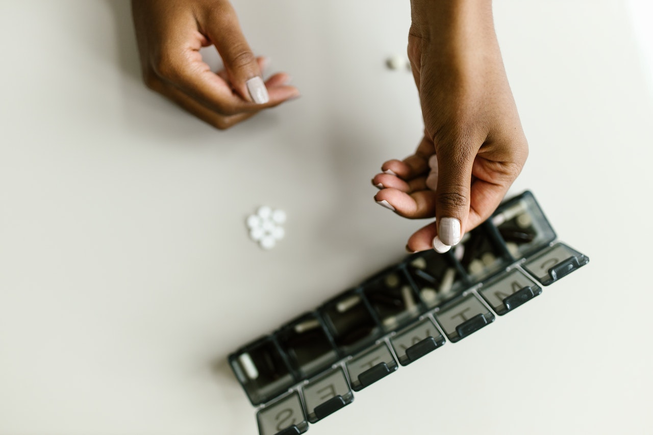 A person with white nail polish is putting a white tablet on a black medicine organizer on top of a white surface