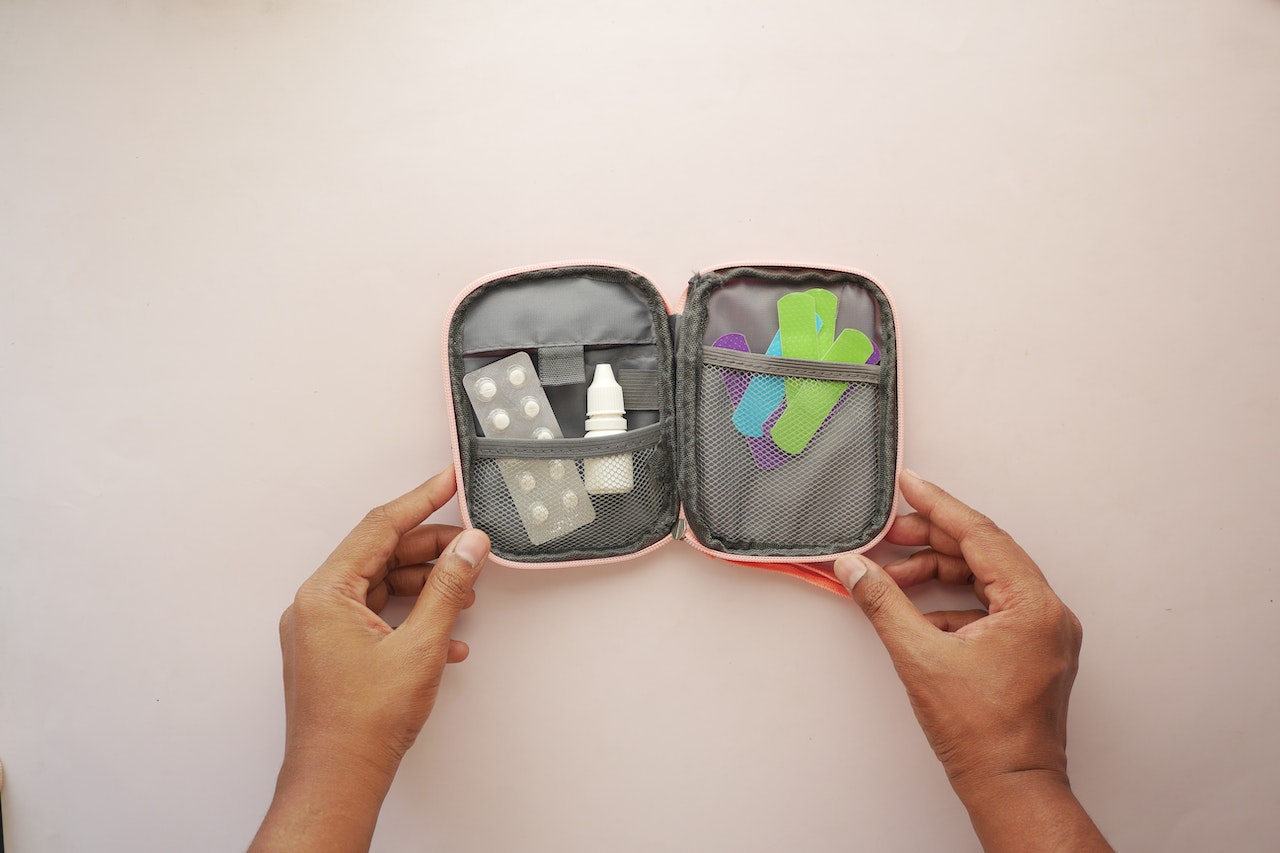 A person holding a gray travel medicine bag with white medicine tablets, a white bottle, and green, violet, and blue colored adhesive bandage on top of a white surface