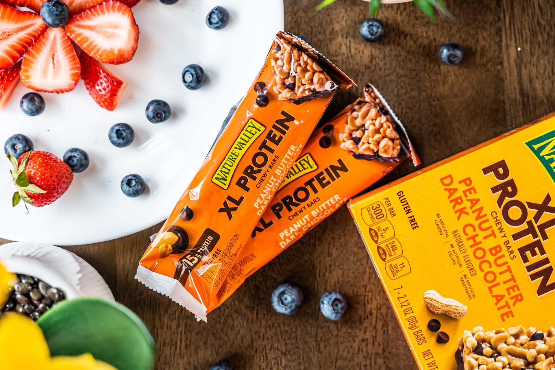 Two XL protein bars with orange packaging near a white plate and a yellow carton box on top of a brown wooden surface