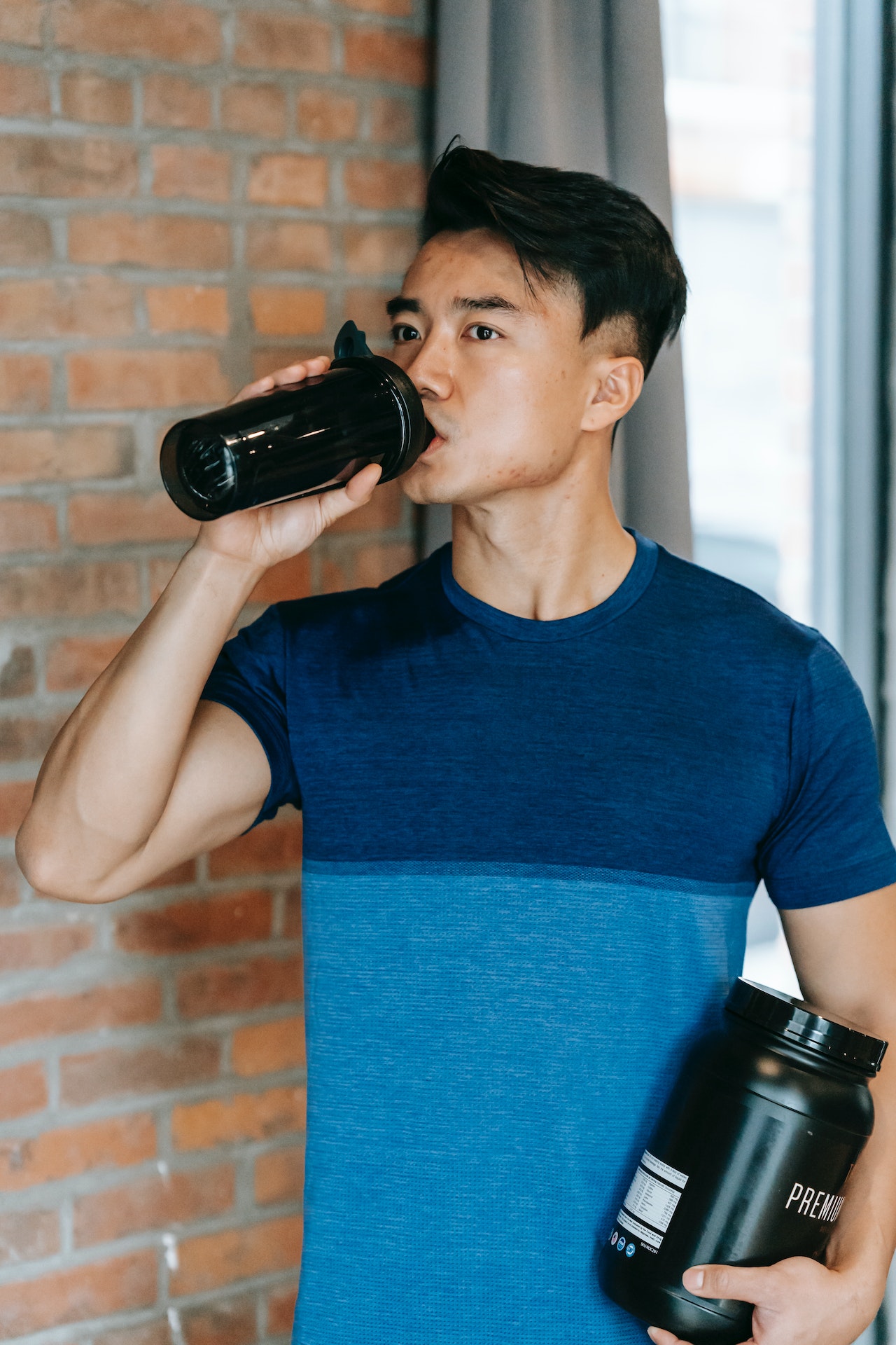 A man wearing a blue shirt is drinking from a black tumbler while holding a black container near a brick wall