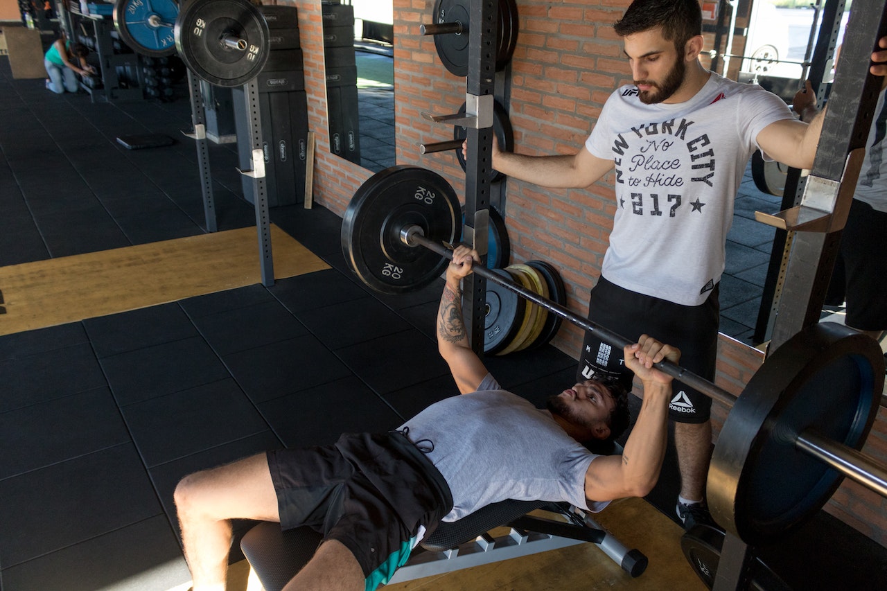 A man wearing a gray shirt and black pants is spotting for the guy with a plain gray shirt and black shorts while using the barbel in the gym