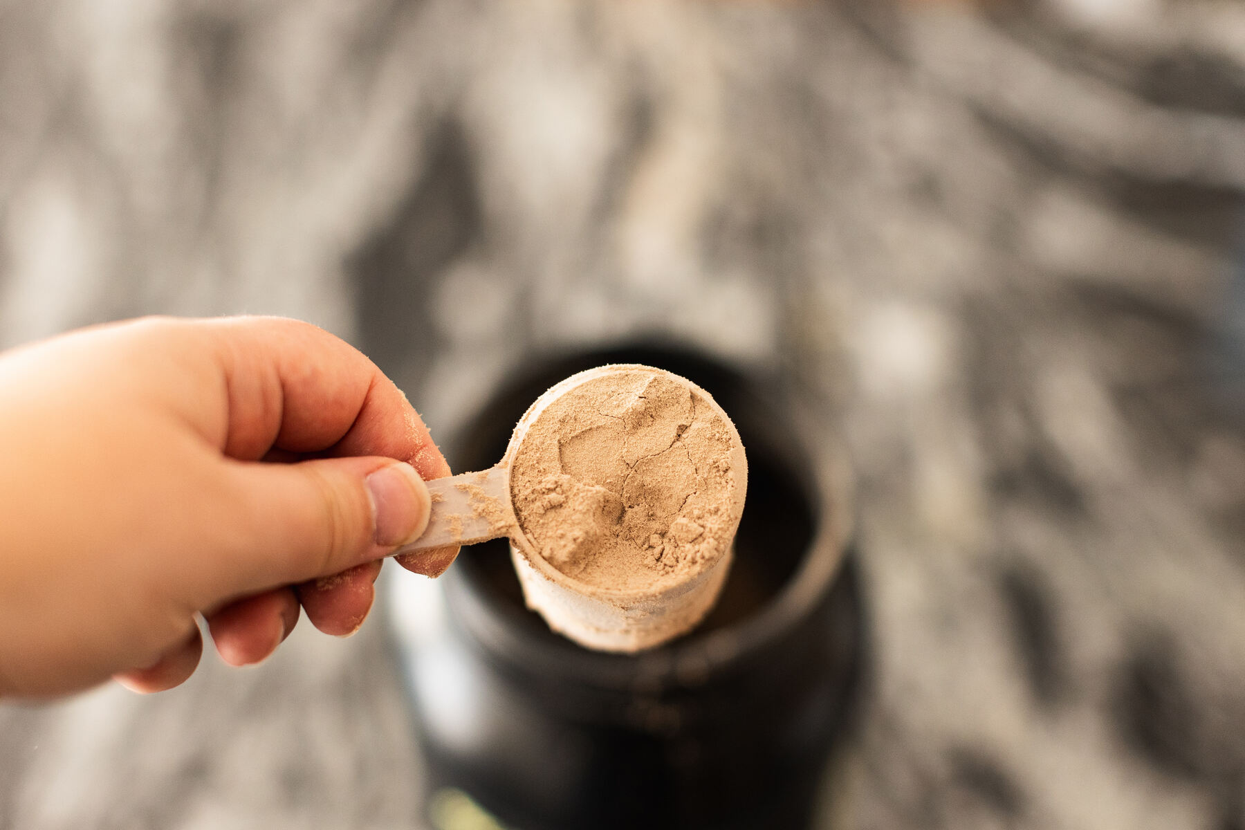 Person holding chocolate whey protein powder in a measuring scoop above a kitchen counter