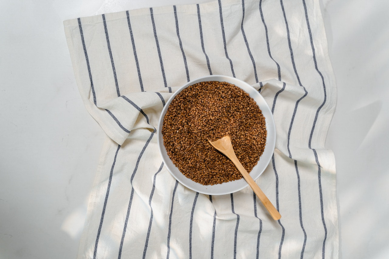 Buckwheat with a brown wooden spoon on a white ceramic bowl placed on top of a striped kitchen towel