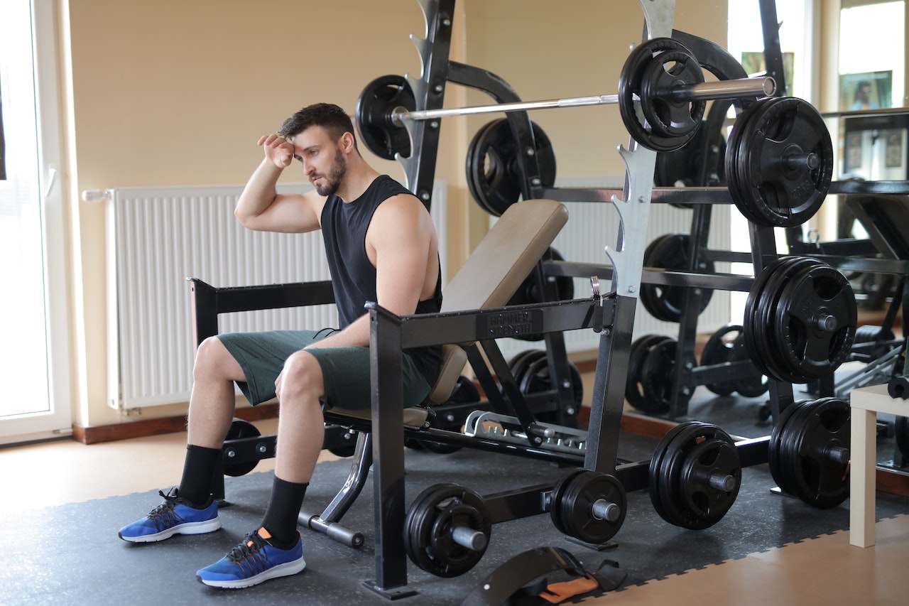 A tired male athlete wearing a black tank top, gray shorts, and blue rubber shoes while sitting on a gym equipment