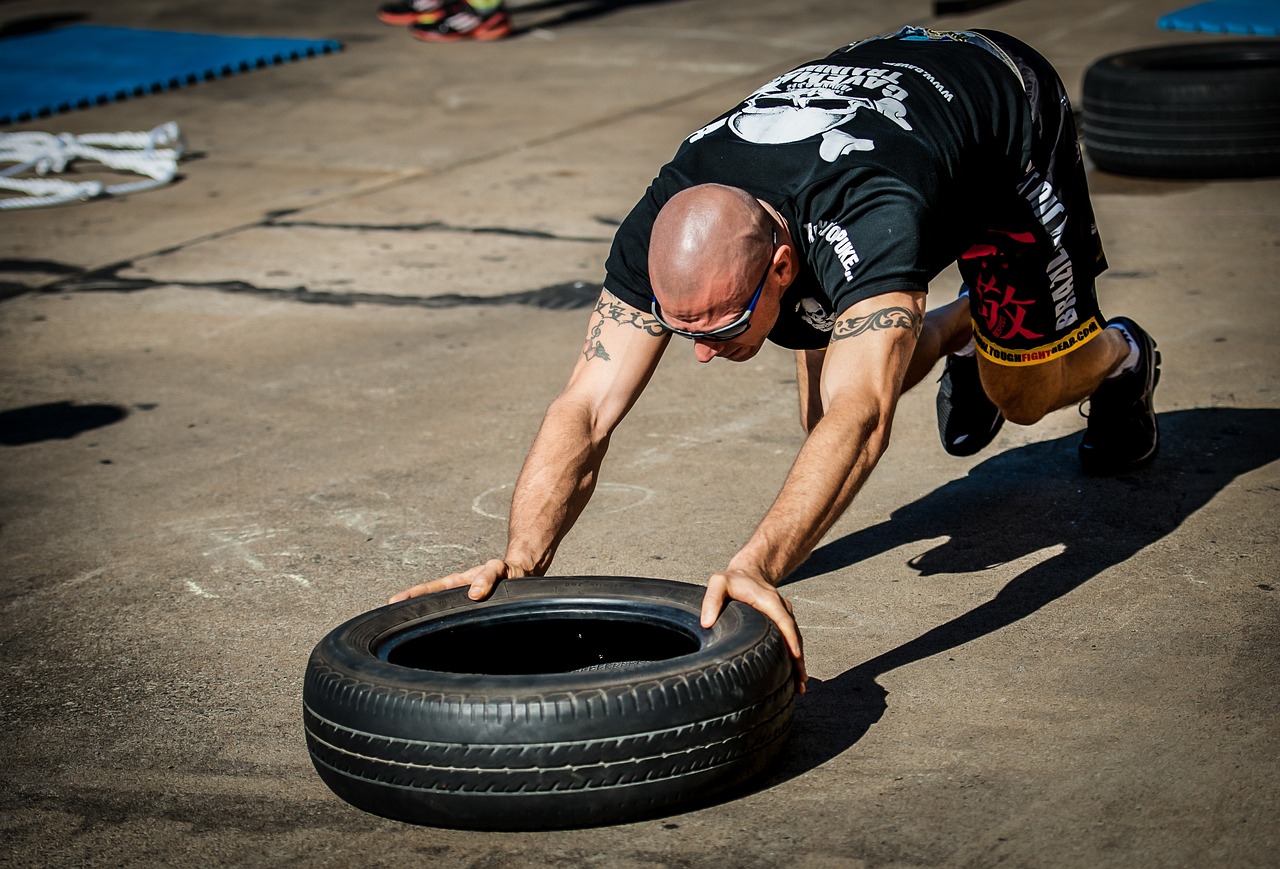 A man wearing a black shirt and black pants is pushing a big black tire on a concrete floor