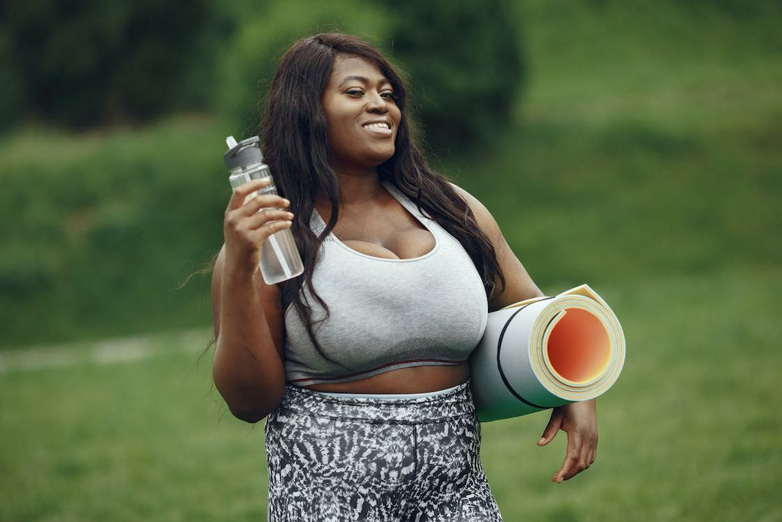 Woman wearing a sports bra and leggings while holding a water bottle and yoga mat in each hands