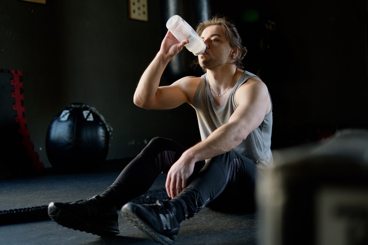 A man wearing a gray tank top, black leggings, and black shoes is drinking from a plastic tumbler while sitting on the gym floor
