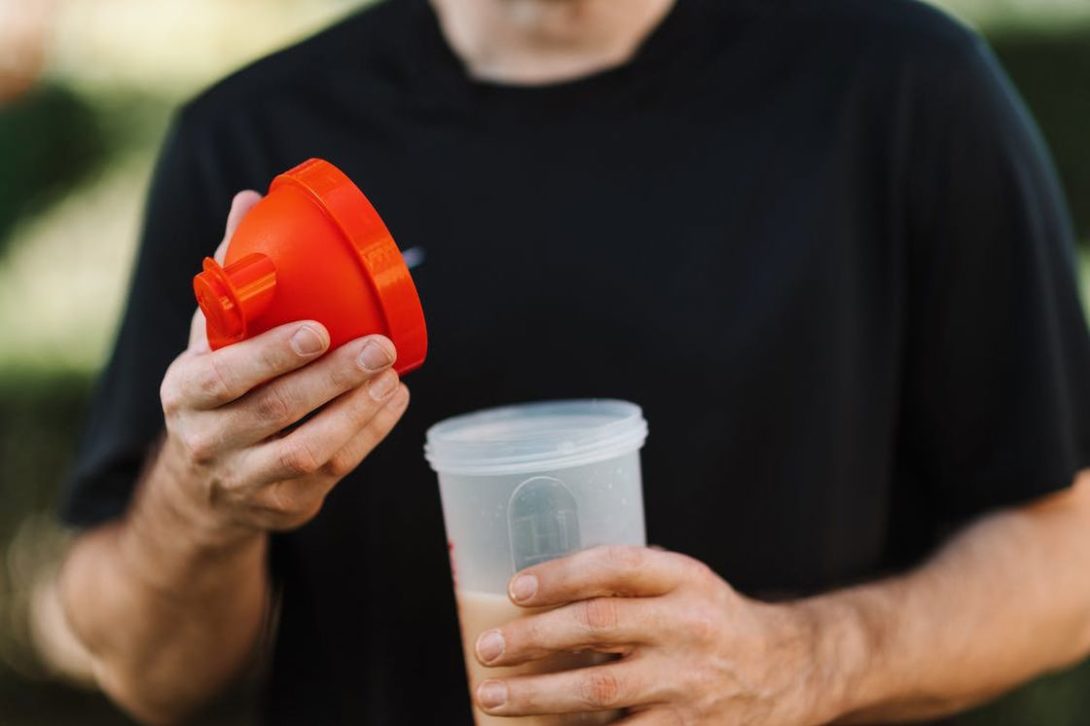 Man wearing a black shirt with a plastic tumbler filled with creatine shake and a red cap