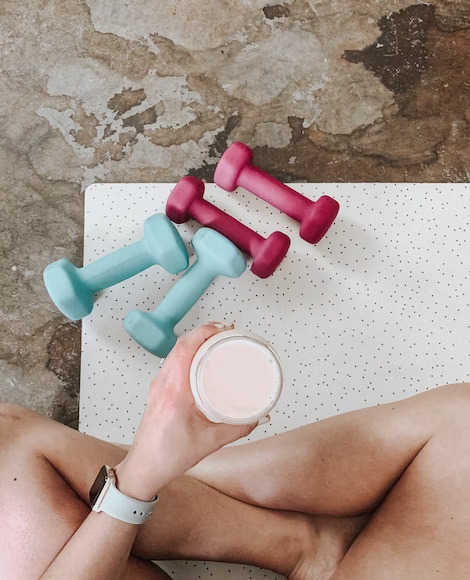 Woman holding a glass filled with supplement while sitting down on a yoga mat with a pair of dumbbells