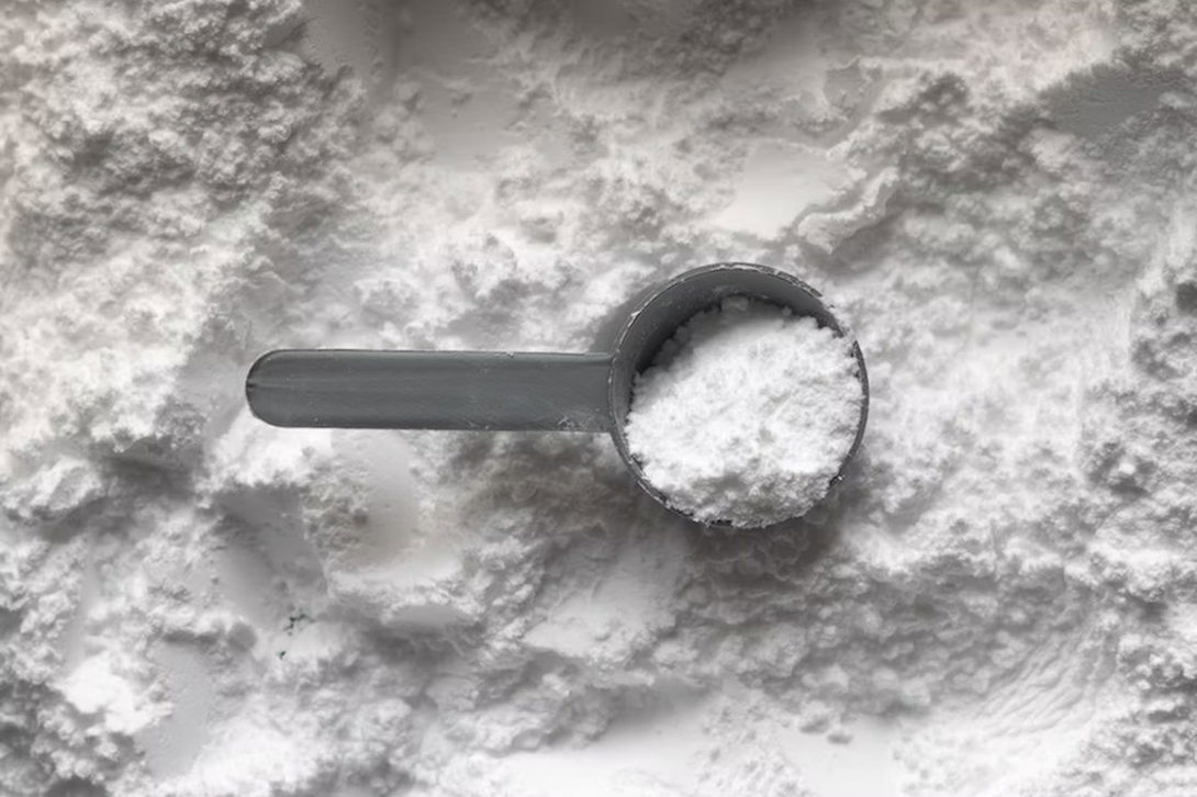 Gray scooper filled with white supplement powder while it's on top of the entire content of white powder