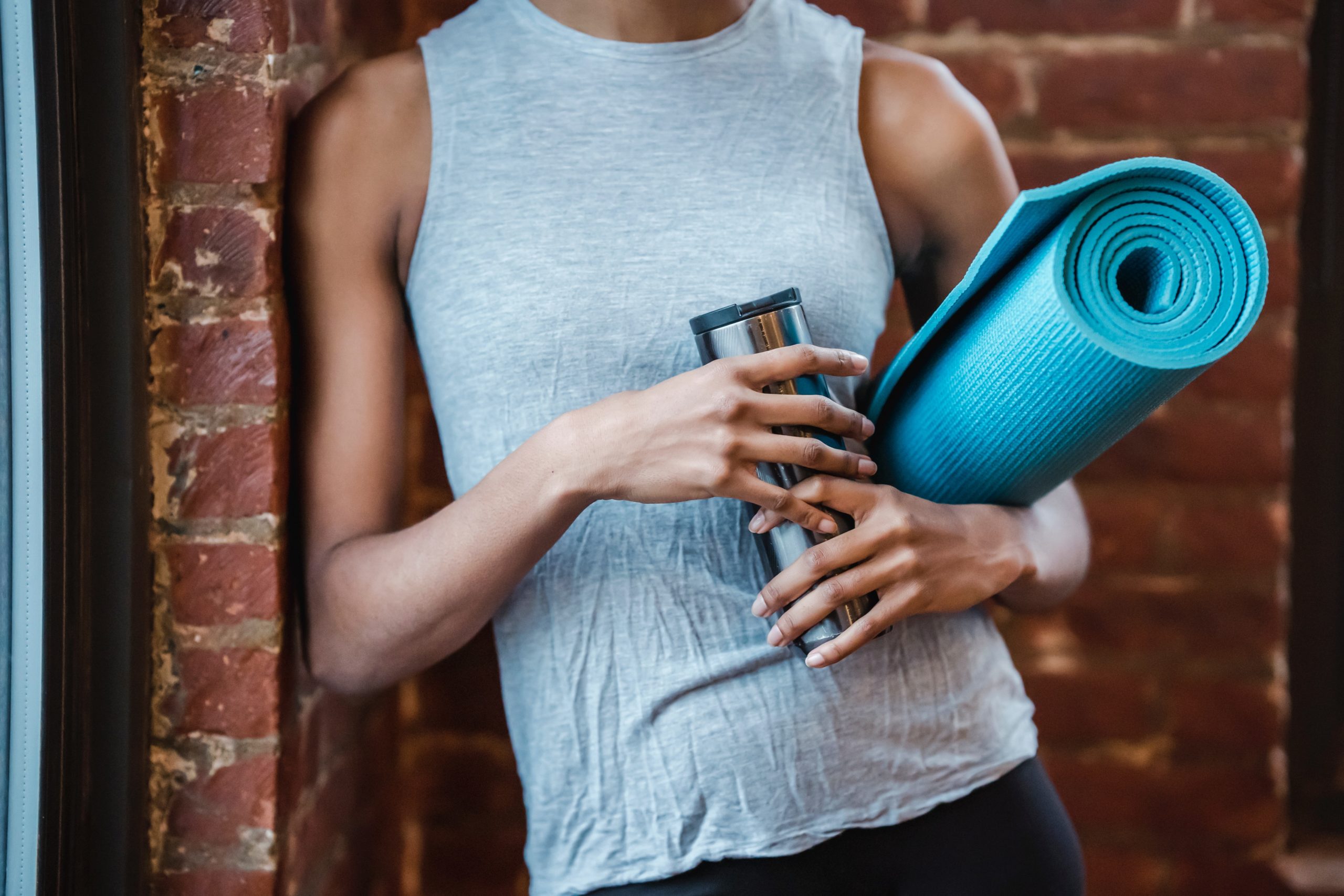 A woman in a gym attire holding an aluminum tumbler and yoga mat on her side