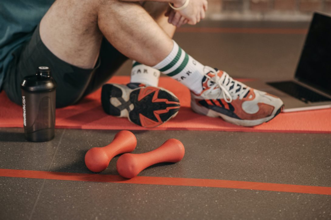 A man sitting with her legs crossed on a yoga mat with a tumbler and two small dumbbells on her side