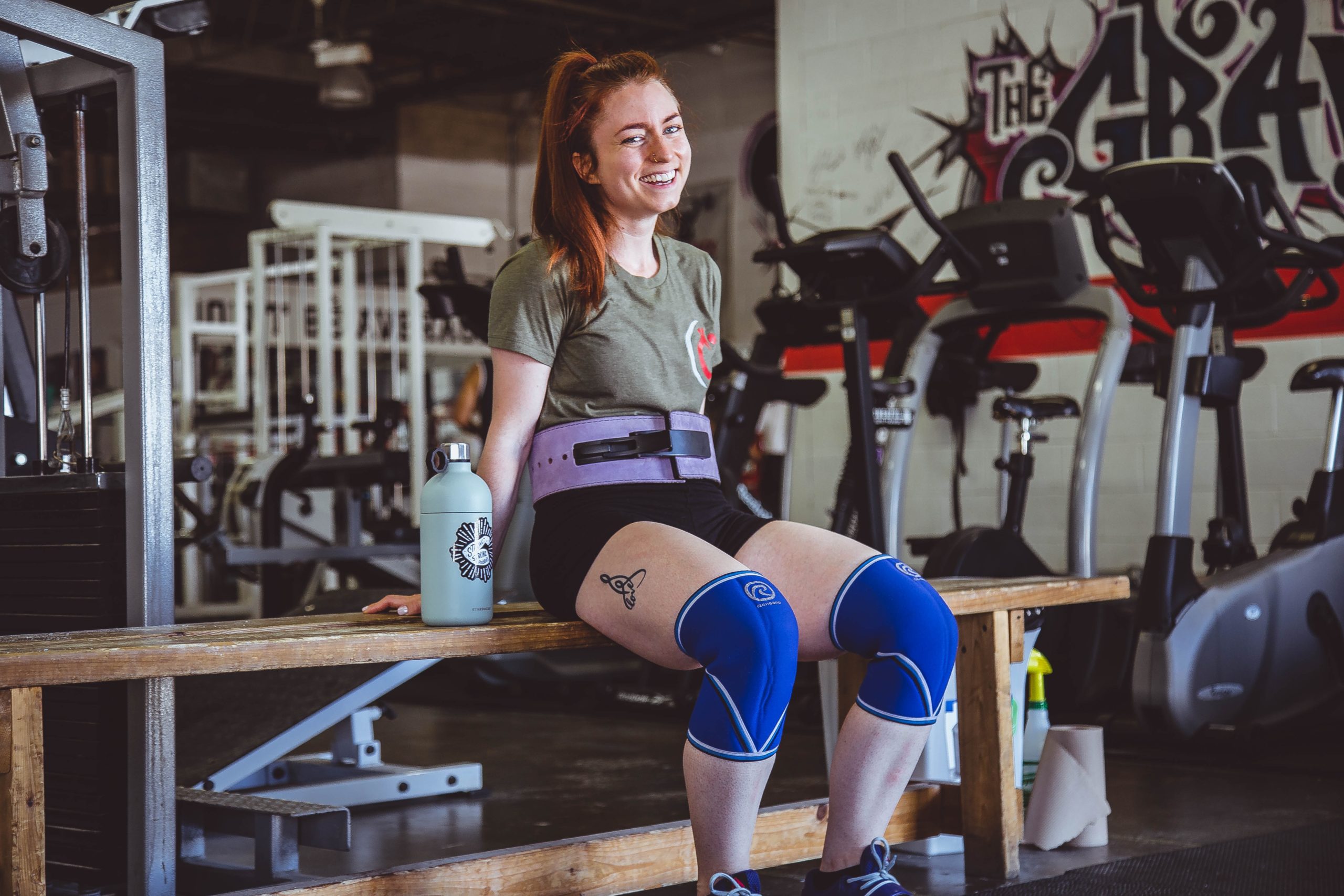 A woman sitting on a wooden bench on the gym with a blue tumbler on her side