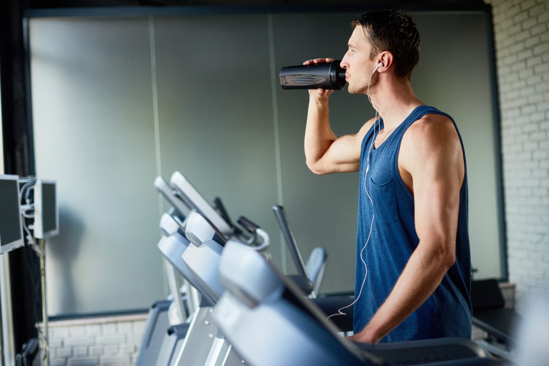 Man in a blue tank top with earbuds walking on a treadmill while drinking from his water bottle
