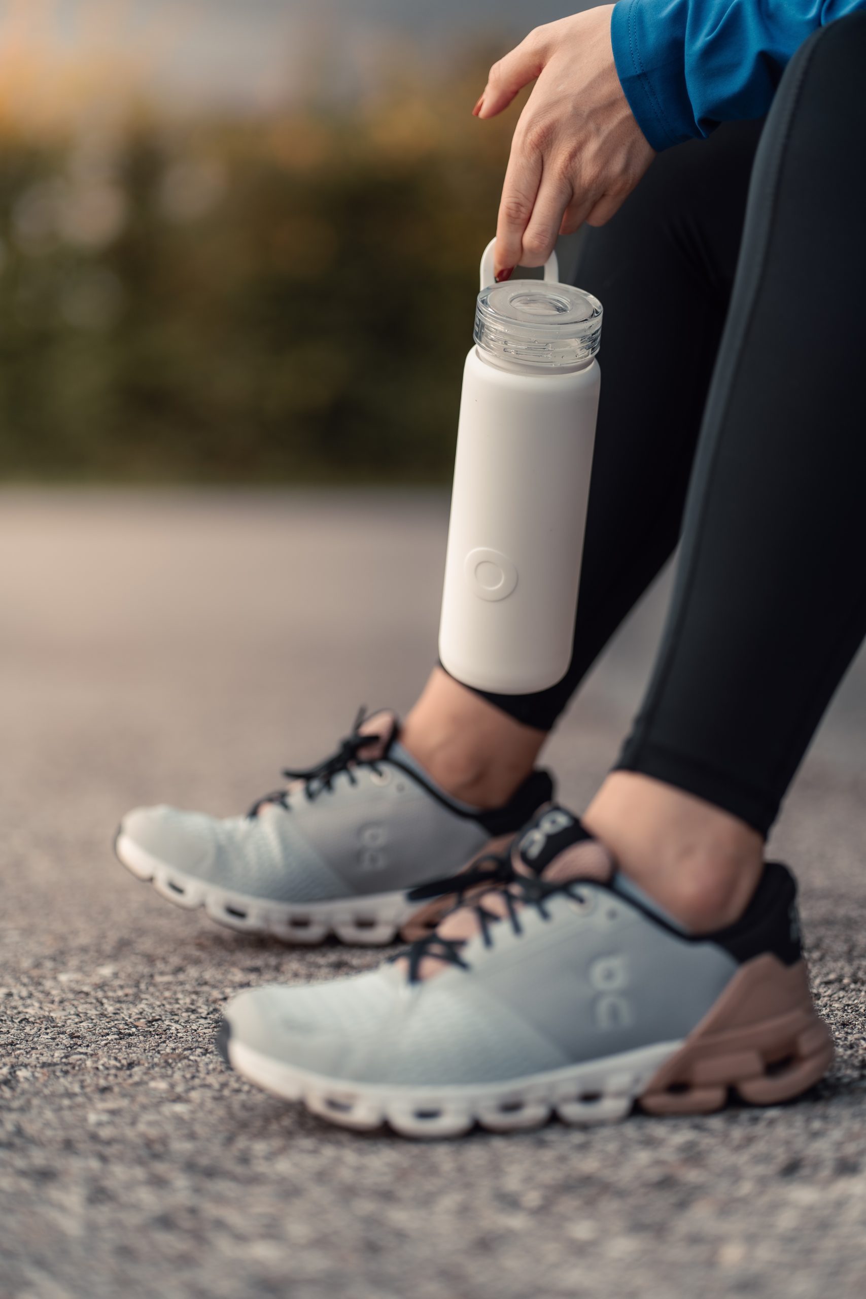 Person holding a white water bottle down by their gray running shoes outside