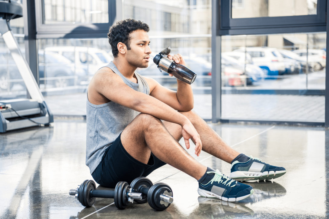 Man sitting on the gym floor drinking pre workout from his gray water bottle