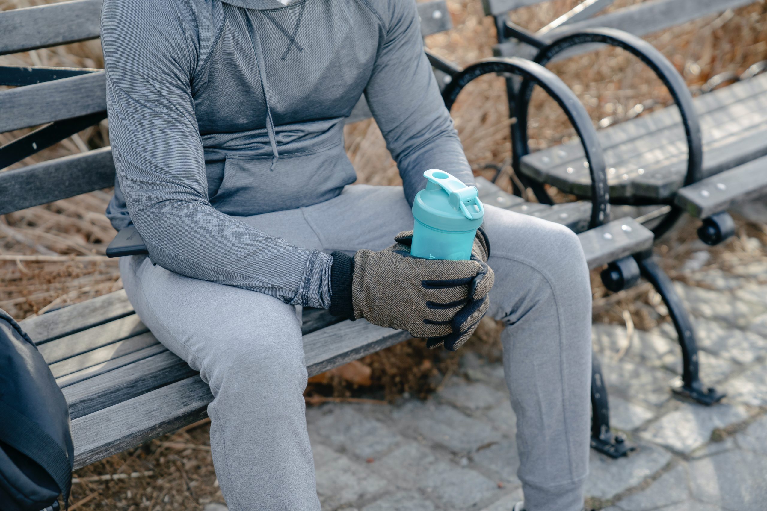 Person in a gray sweatsuit sitting on a park bench holding a light blue water bottle