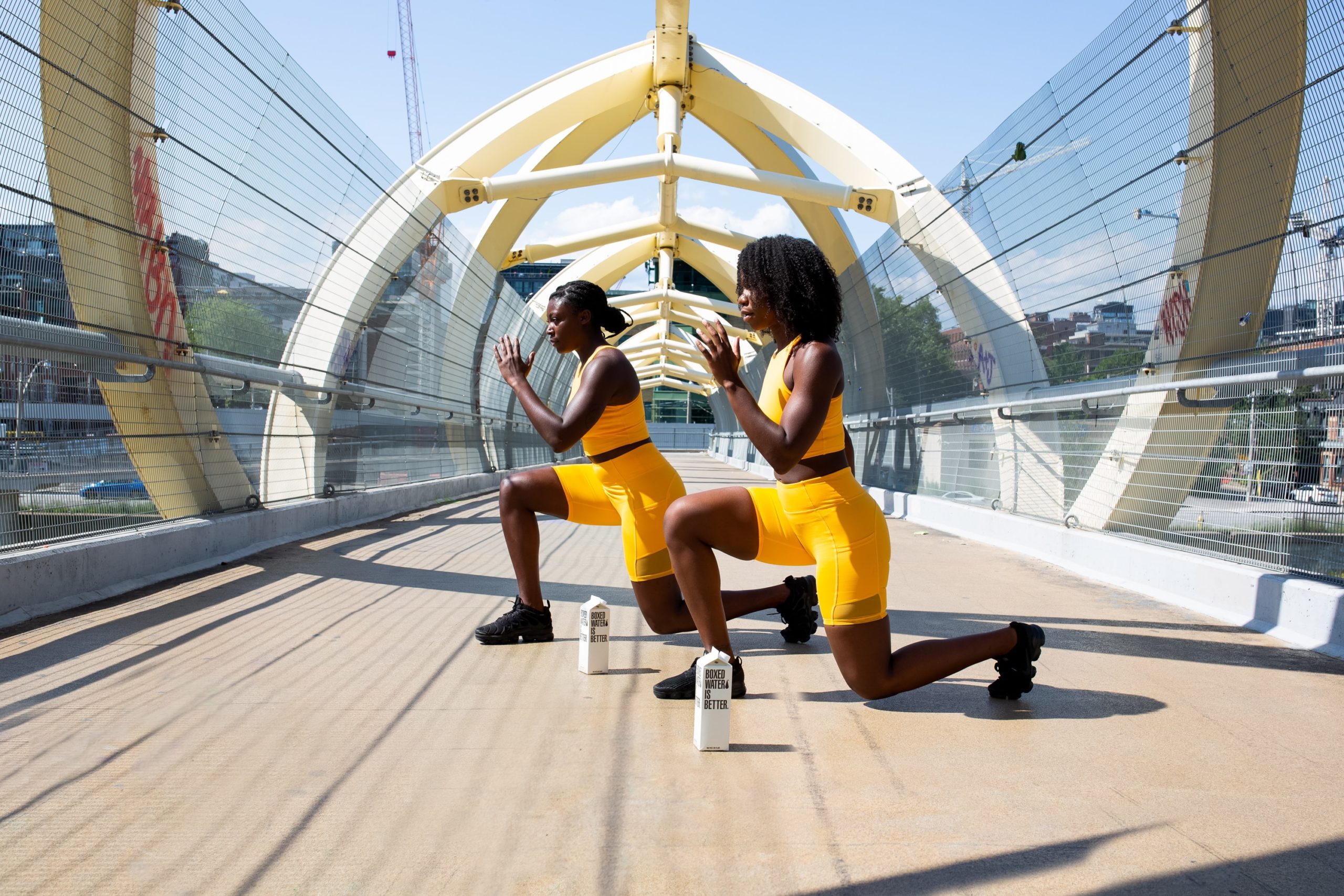 Two woman wearing bright yellow gym clothing exercising on a bridge with two boxed water beside them