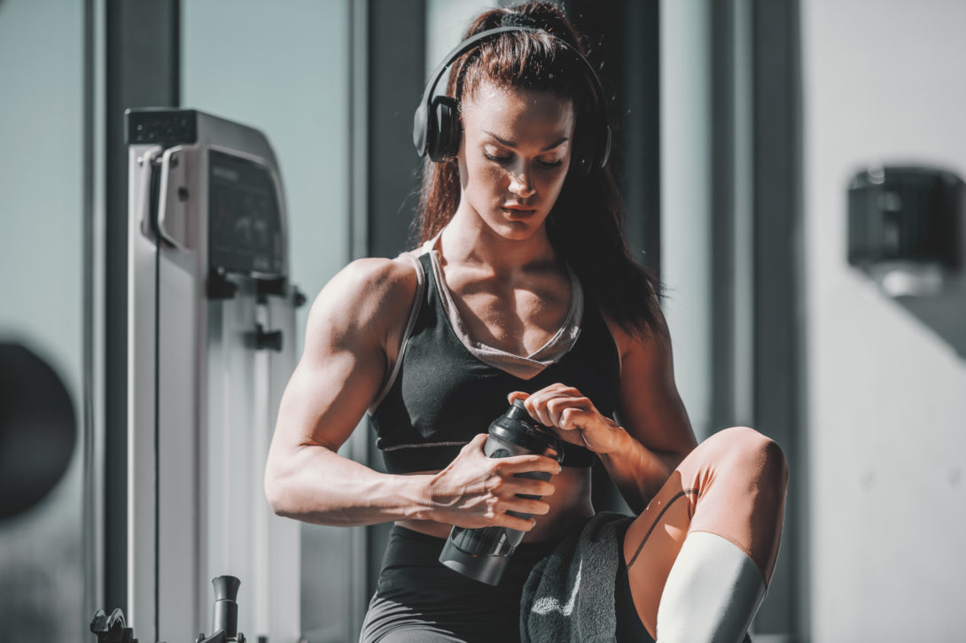 Strong muscular female bodybuilder sitting in gym with headphones on holding a water bottle