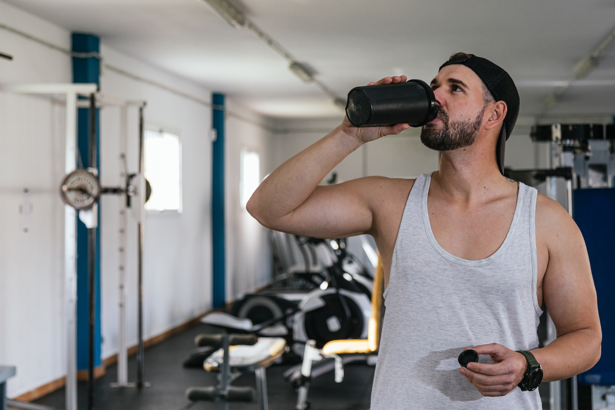 Male in a gray tank top drinking pre workout from a black water bottle at the gym