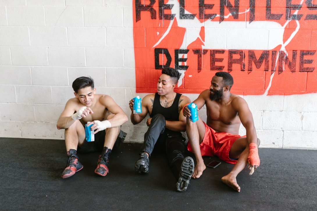 Three men sitting on a gym holding their own beverages while resting