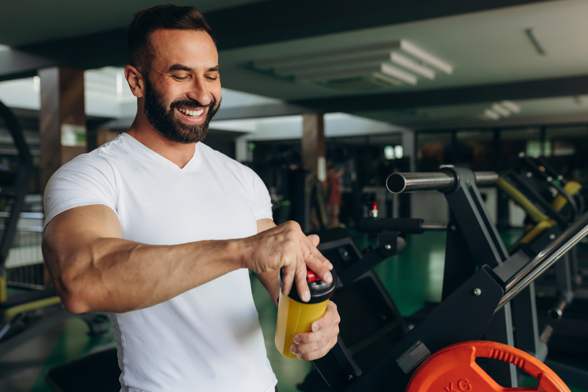 Man smiling in a gym holding a shaker bottle while next to exercise equipment