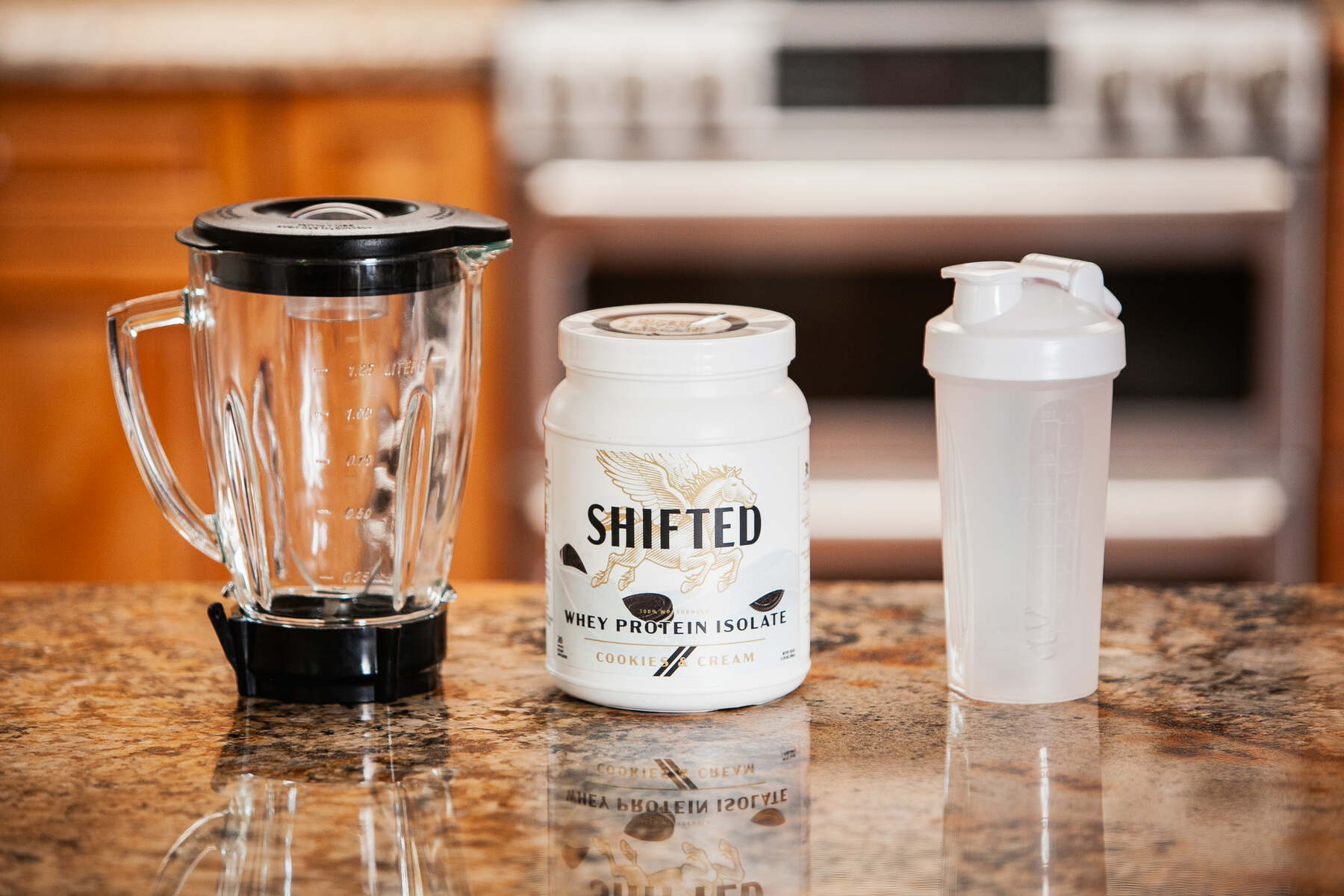 A container of "SHIFTED" Whey Protein Isolate in Cookie & Cream flavor, with a blender and a shaker bottle on a kitchen counter