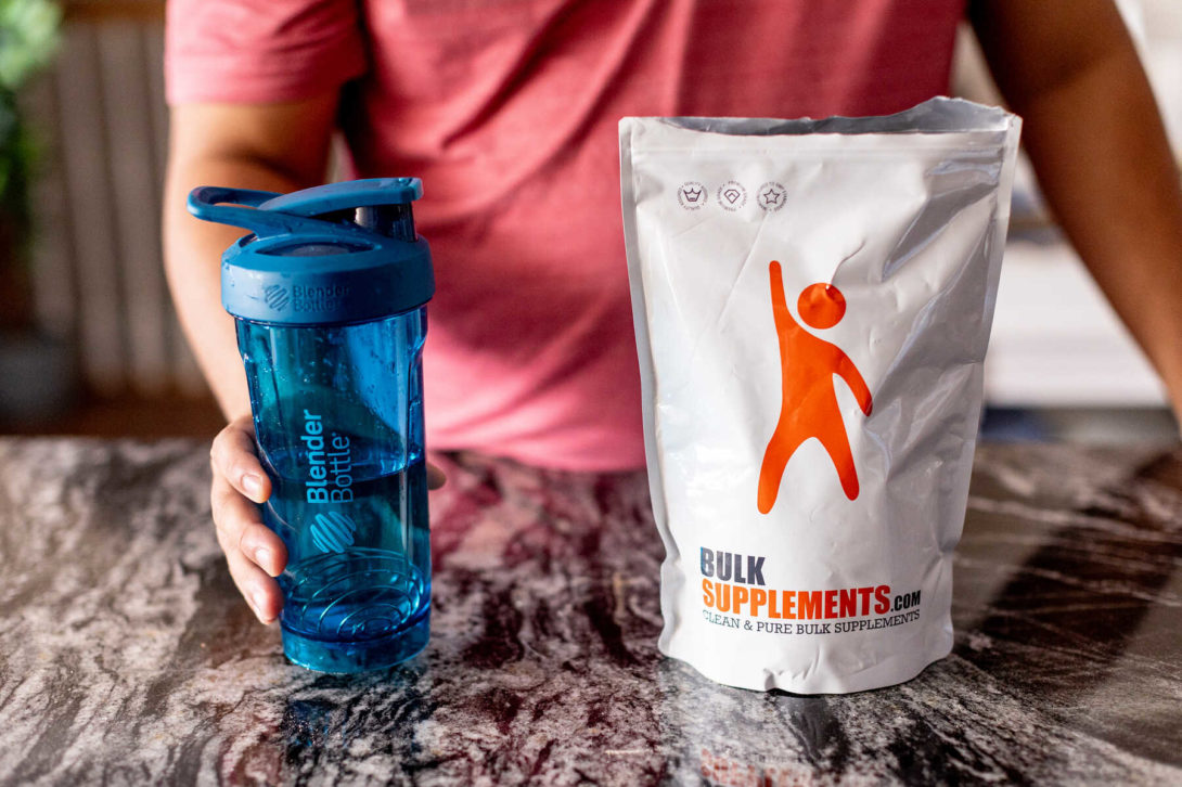 Man holding a blue bottle blender next to a Bulk Supplements bag on a black granite countertop