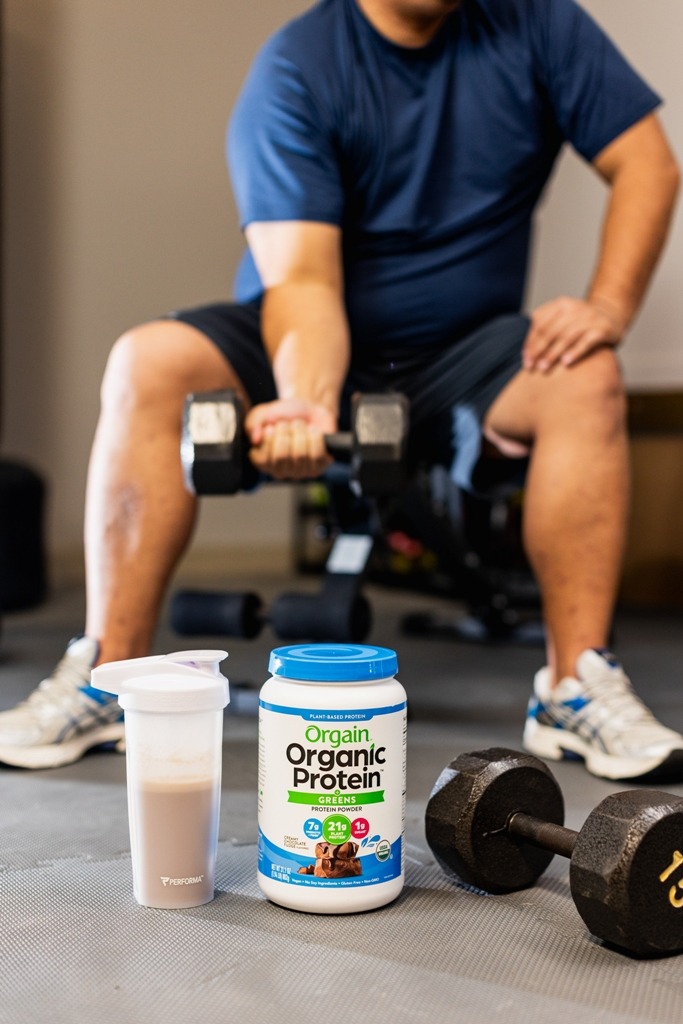 Orgain Protein & Greens Plant-Based Protein Powder beside a dumbbell, water bottle with a man exercising on the background