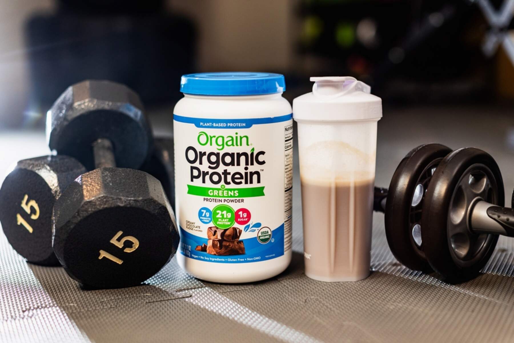 Orgain Protein & Greens Plant-Based Protein Powder bottle beside a pair of dumbbells and a water bottle filled with protein shake