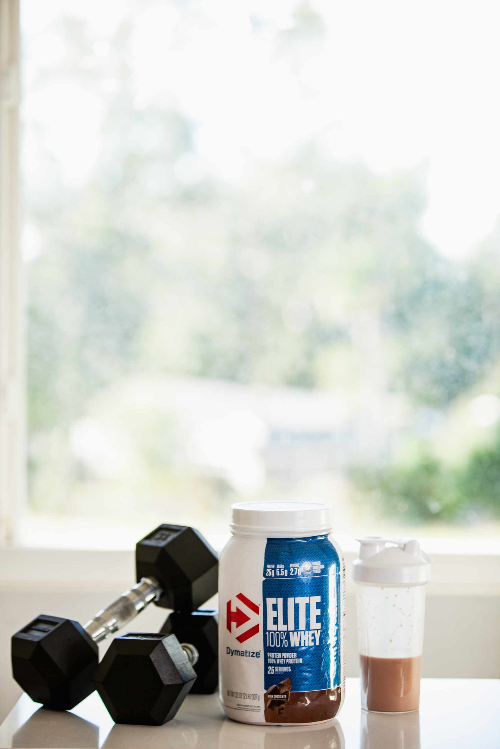 Elite 100% Whey protein powder beside a pair of dumbbells and a water bottle filled with protein shake