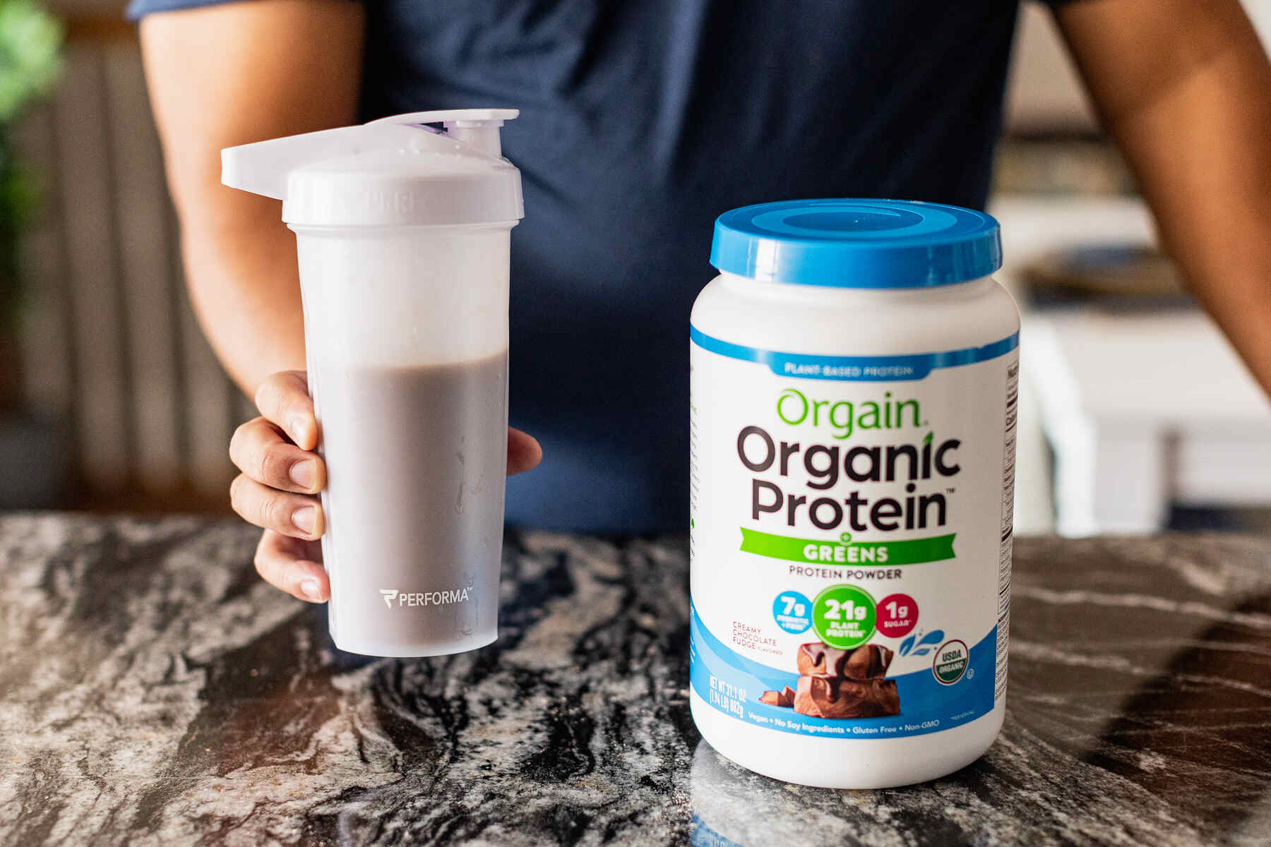 Person holding a protein shake in a water bottle beside a bottle of Orgain Organic Protein powder