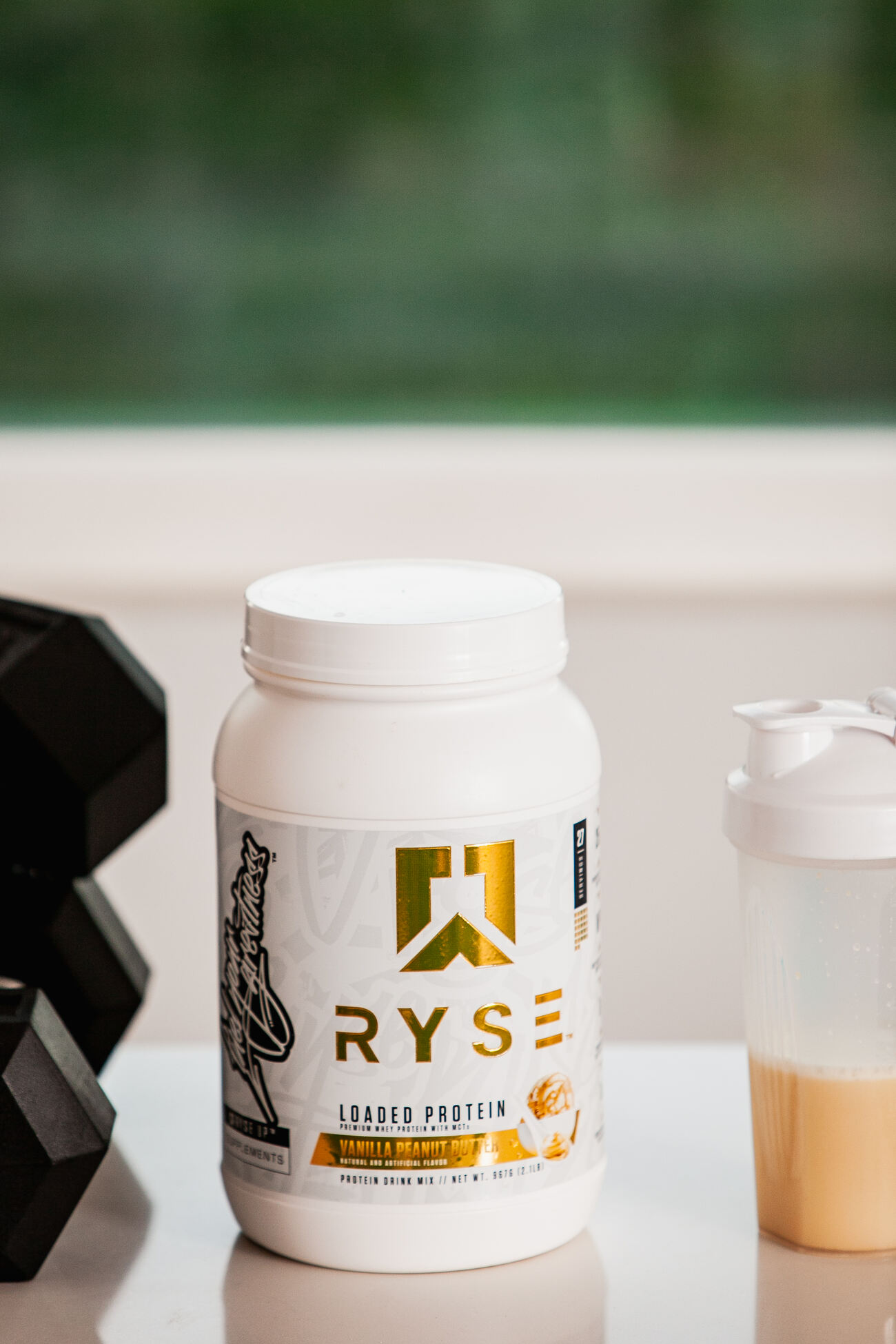 A container of RYSE Loaded Protein in a white bottle with gold and black labeling placed beside a protein shaker on a white surface with a blurred green outdoor background