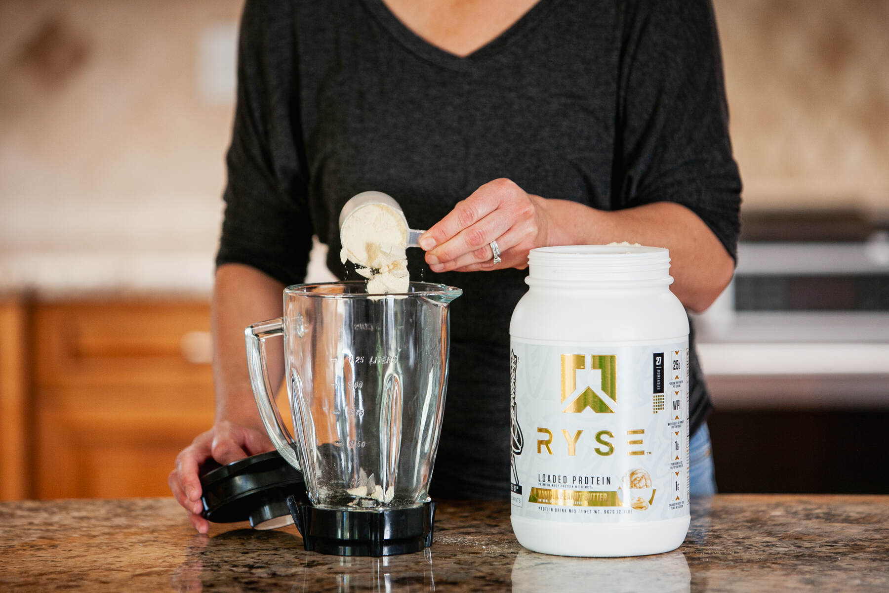 A person in a black top is pouring vanilla peanut butter flavored RYSE Loaded Protein powder into a blender, with the container placed on a kitchen counter