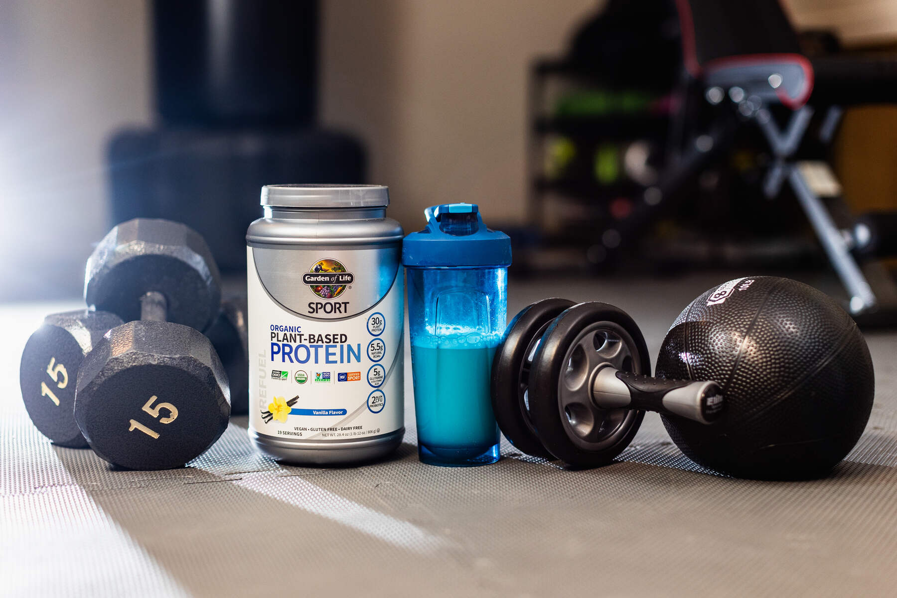 A workout area with a silver container of Garden of Life Sport Organic Plant-Based Protein, a blue protein shaker, dumbbells, and a black exercise ball