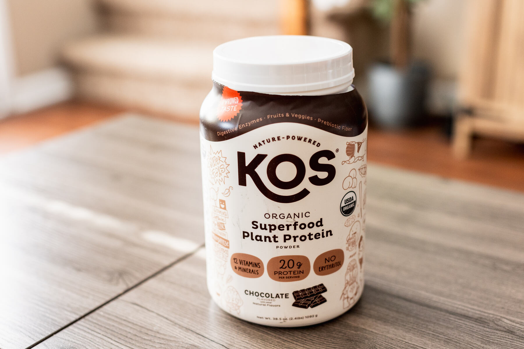 Brown container of KOS Organic Superfood Plant Protein powder on a wooden surface, with a focus on the chocolate flavor