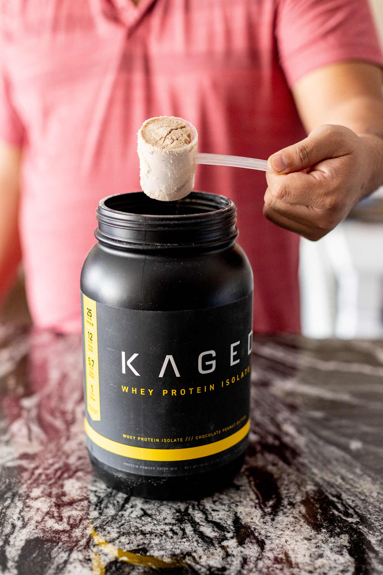 Man holding a scoop of Kaged whey protein powder on a black granite countertop