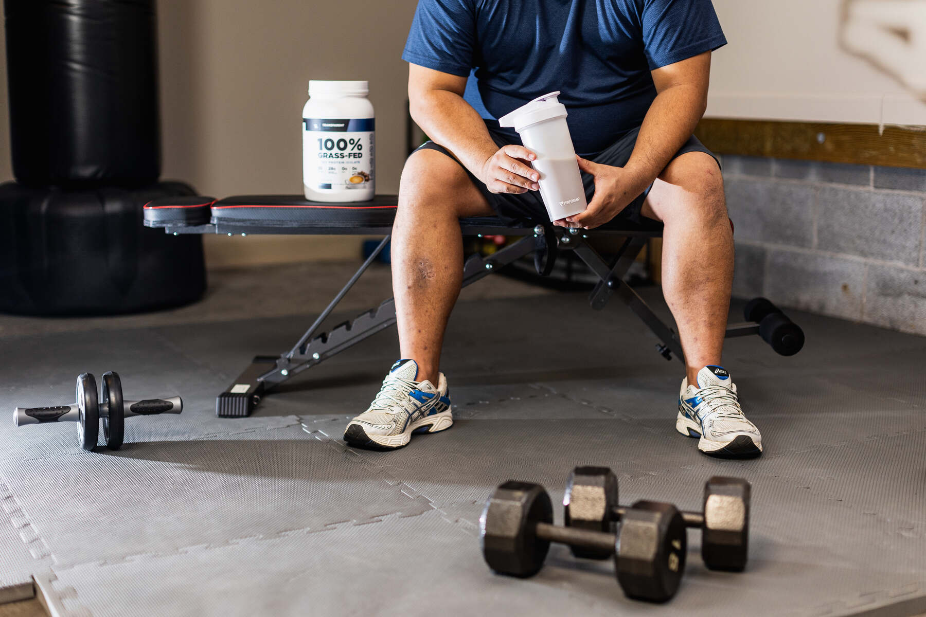 Man holding a protein powder drink while sitting on a workout bench in his garage gym
