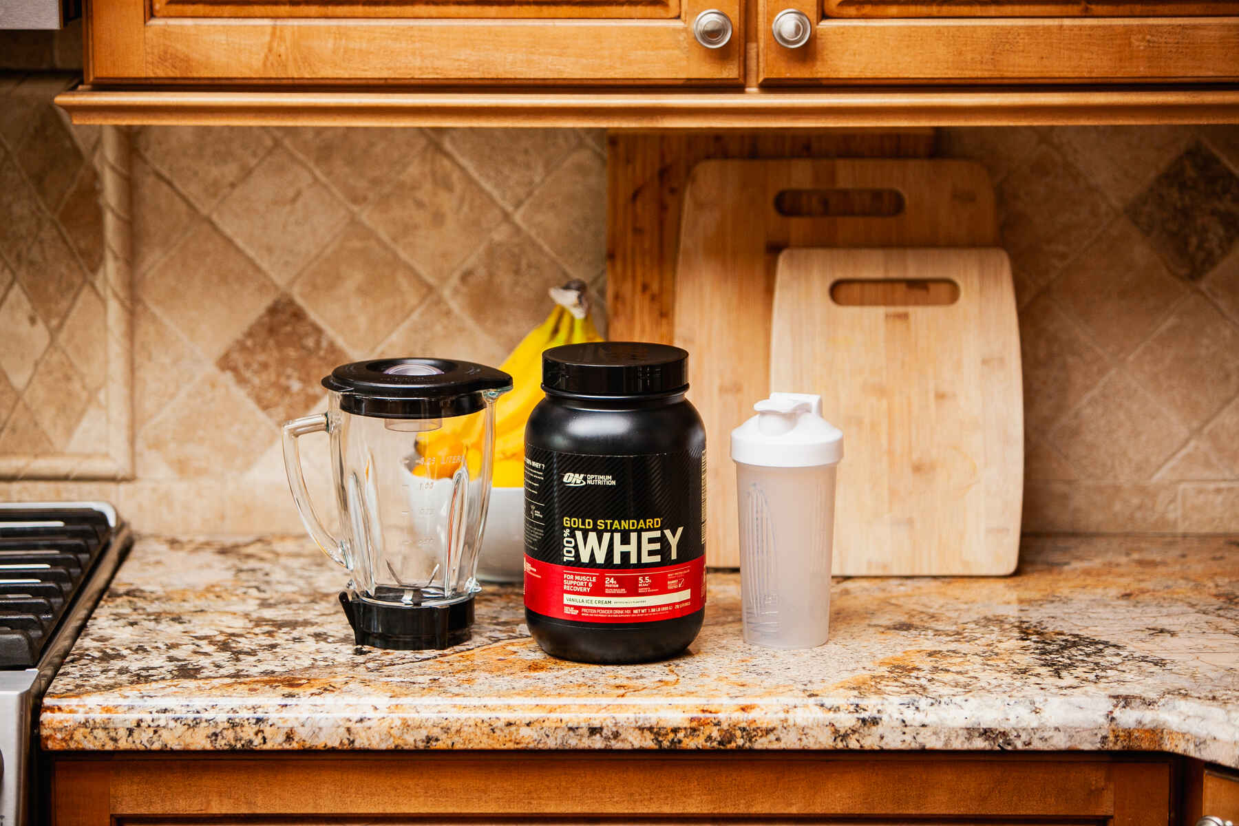 A kitchen counter with a blender, protein powder jar, and a shaker bottle, with wooden cabinets in the background