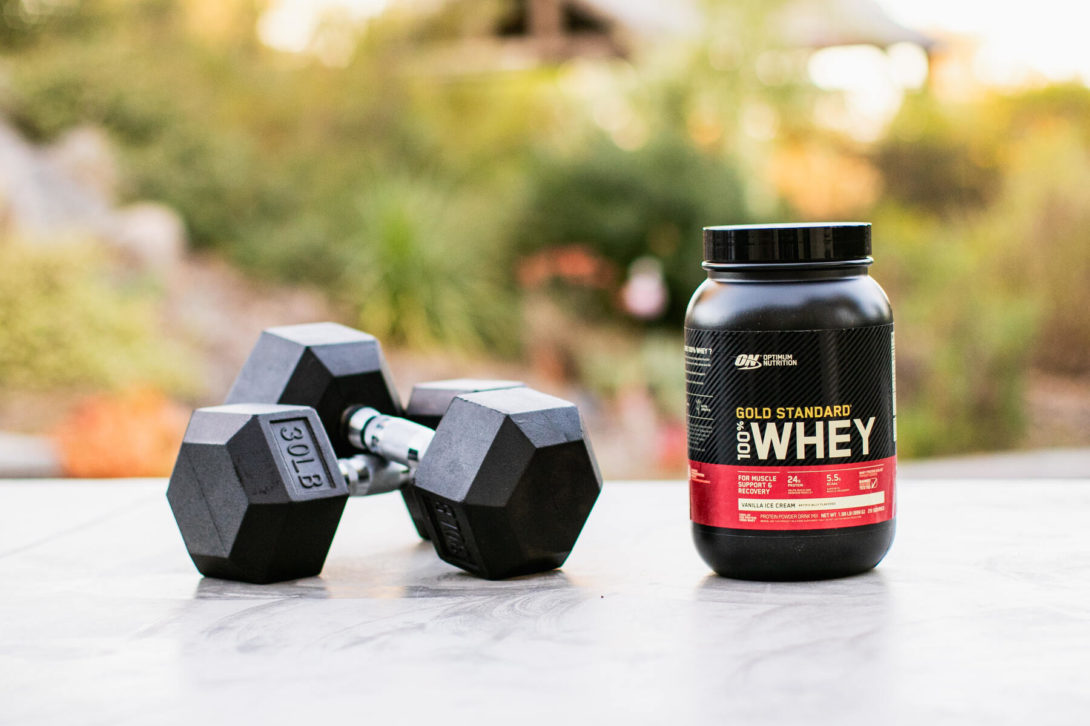 A black dumbbell and a jar of protein powder on an outdoor surface with greenery in the background