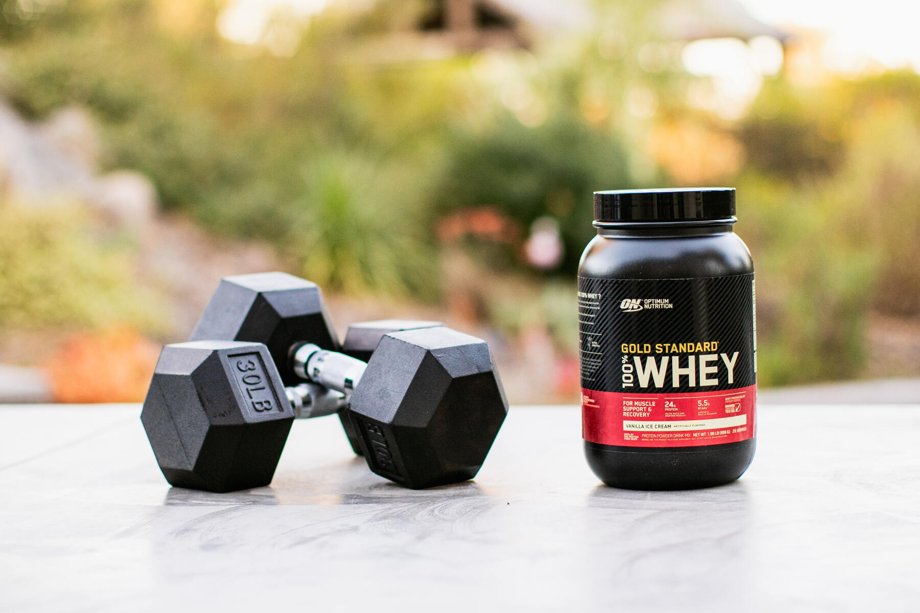 A black dumbbell and a jar of protein powder on an outdoor surface with greenery in the background