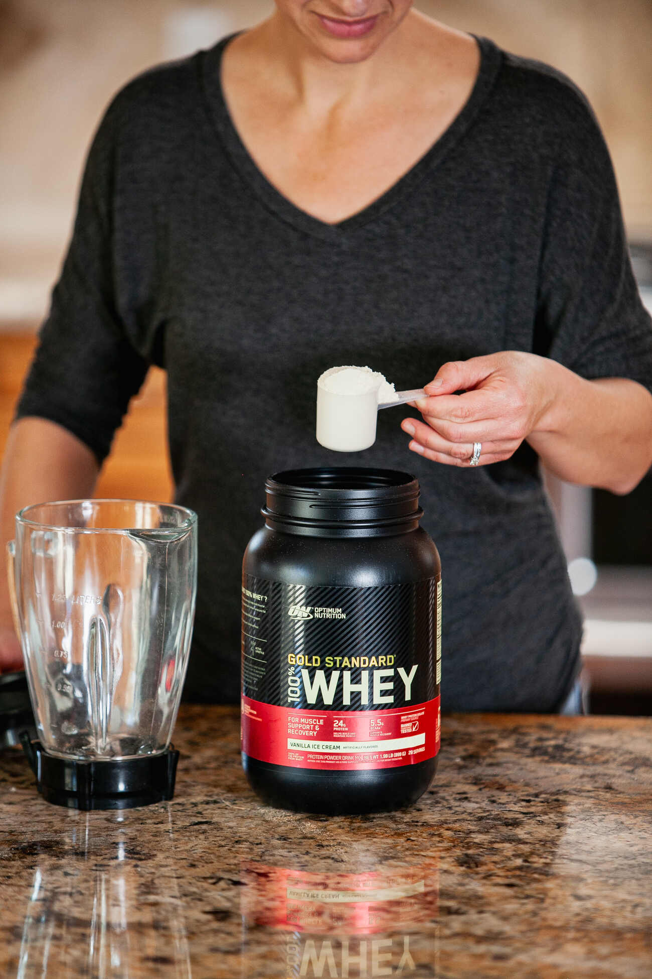 A woman holding a scoop of protein powder above a jar, with a blender on the kitchen counter