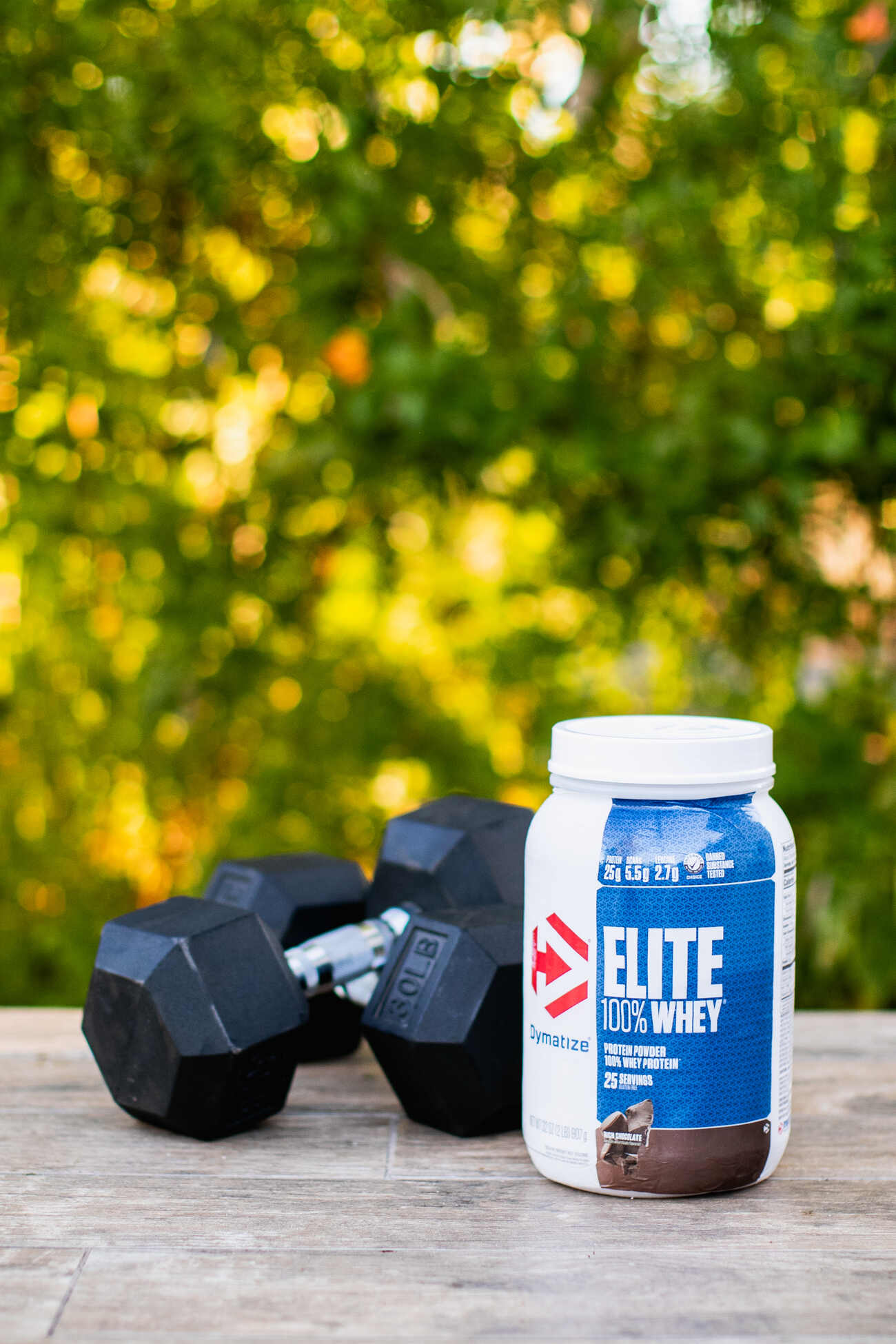 A protein powder jar next to dumbbells on a wooden surface outdoors