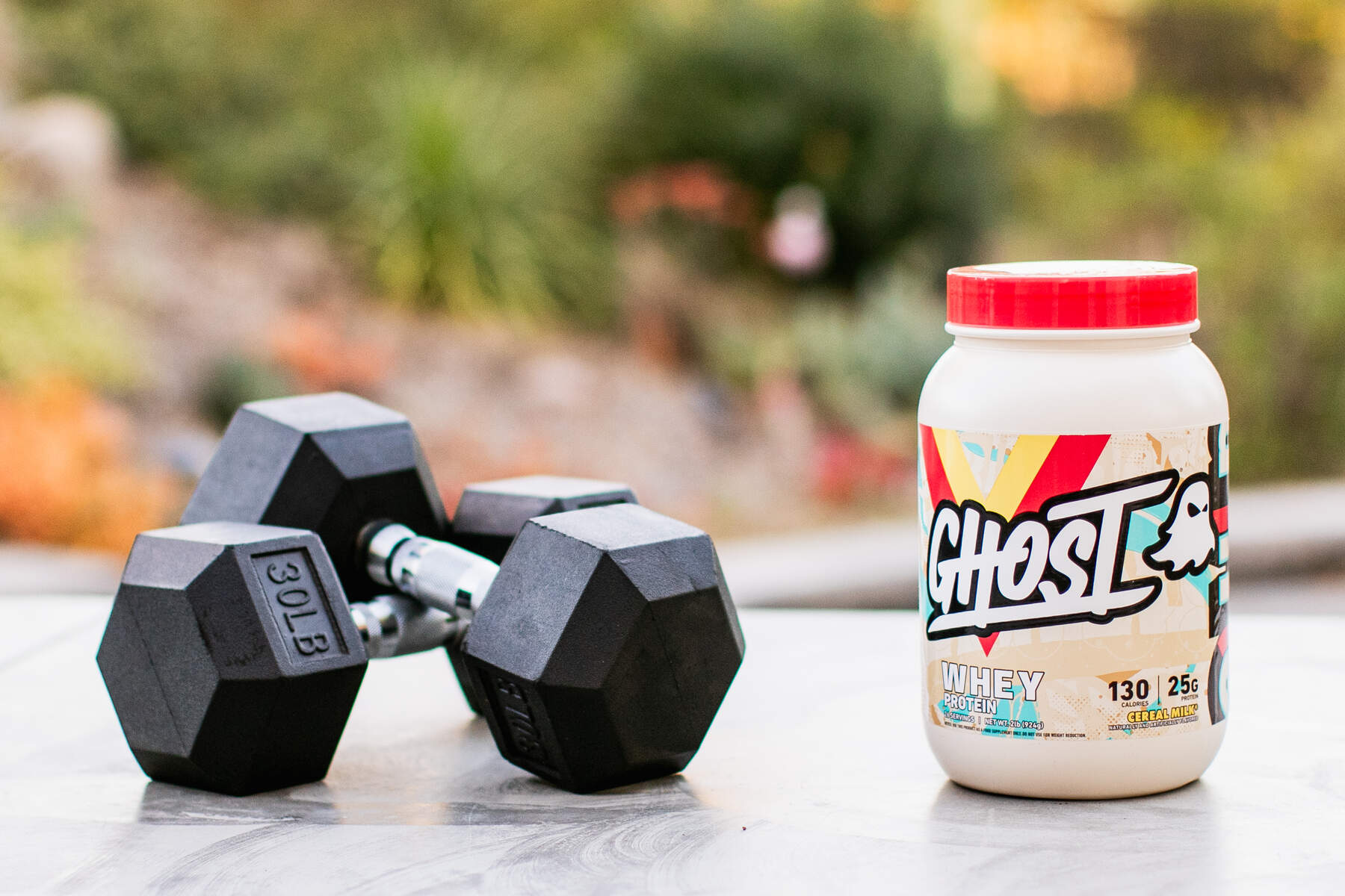 30lb metal dumbbells and Ghost whey protein supplement outdoors on a concrete table